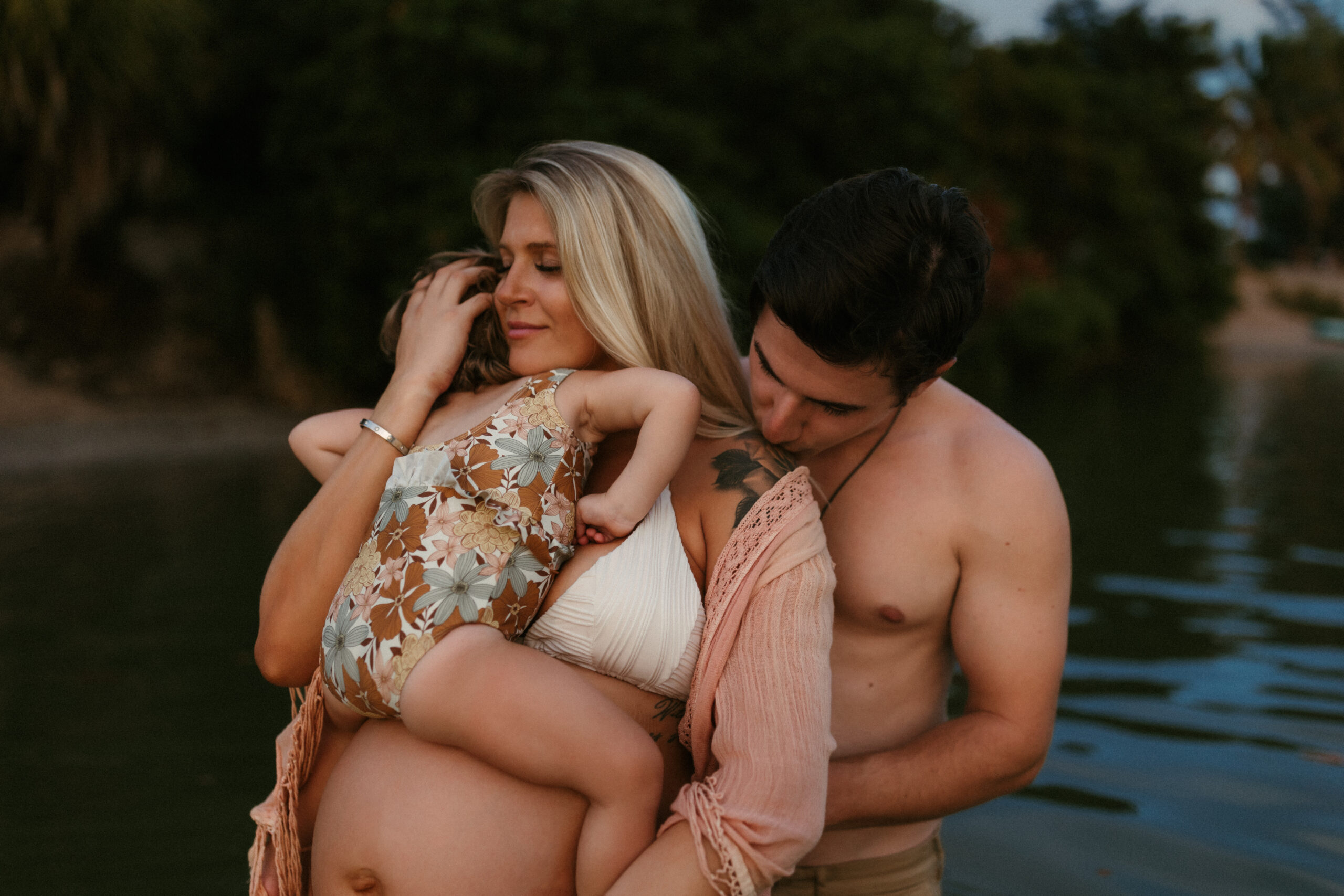 pregnant mother holding her young daughter on her belly while her husband is holding her close and kissing her on the shoulder